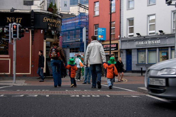 2 children with green hats crossing the road with parent. 